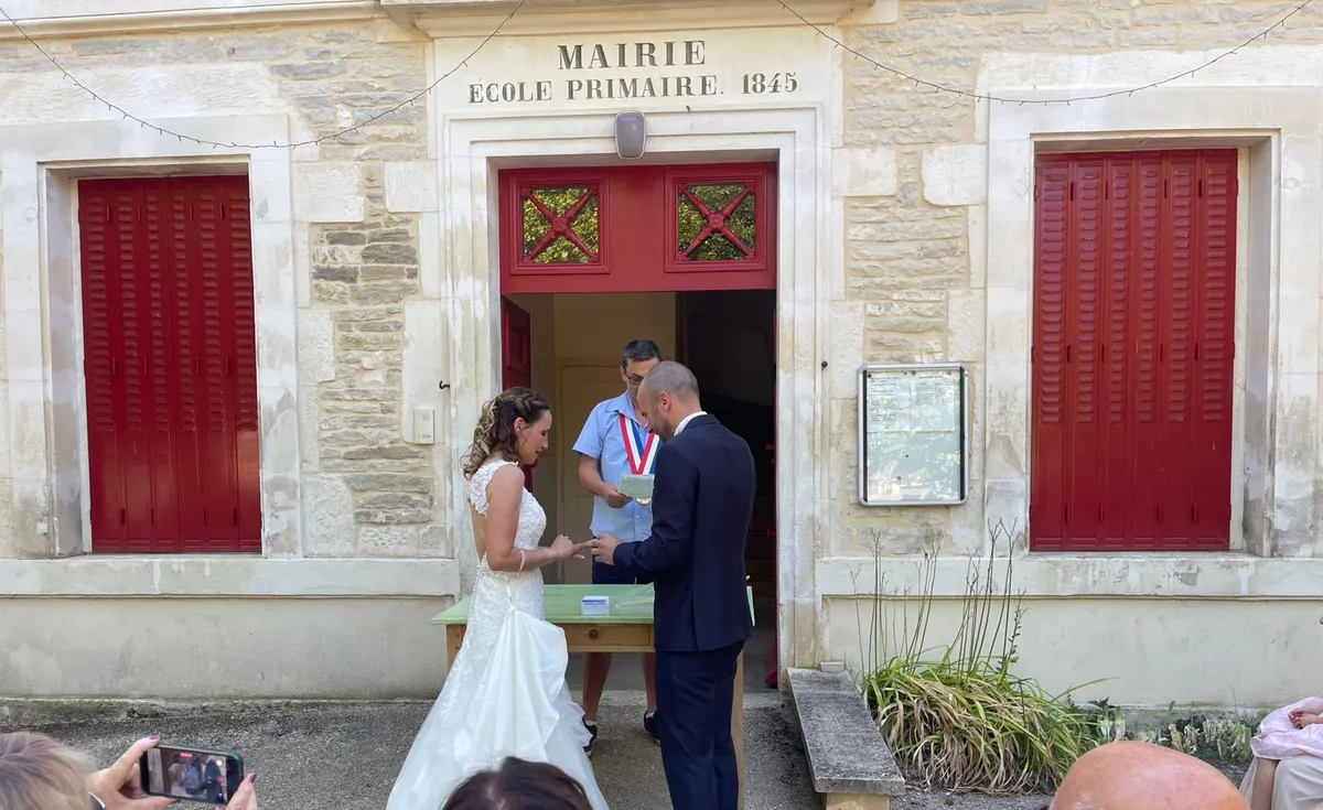 Les mariés échangent leurs alliances devant la mairie, en présence du maire ceint de son écharpe.