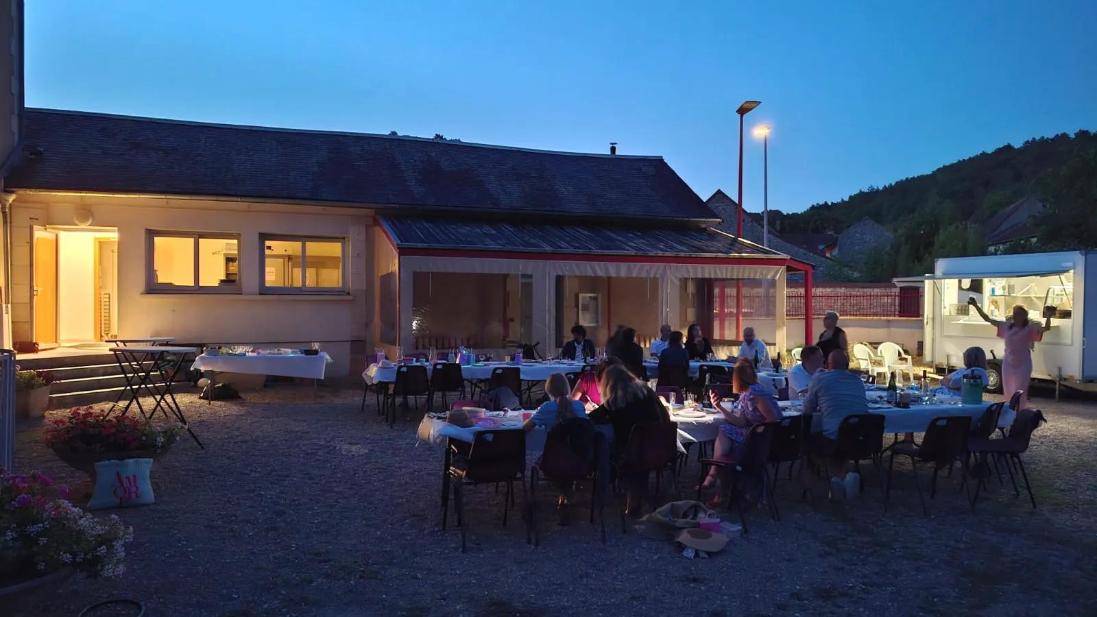 La grande tablée réunie à la tombée de la nuit, bâtiment et food truck éclairés.